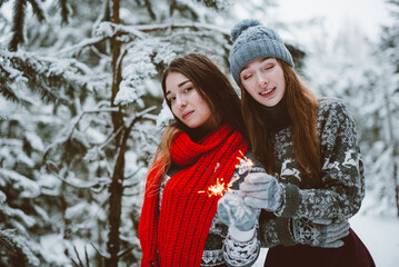 Fototapeta na wymiar Close up fashion portrait of two sisters hugs and having fun, holding sparklers in winter time forest, wearing sweaters and scarfs,best friends couple outdoors, snowy weather