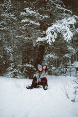 Fototapeta na wymiar Close up fashion portrait of two sisters hugs and having fun, ride on sled in winter time forest, wearing sweaters and scarfs,best friends couple outdoors, snowy weather