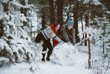 Close up fashion portrait of two sisters hugs and having fun, make snowman in winter time forest, wearing sweaters and scarfs,best friends couple outdoors, snowy weather