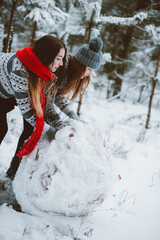 Close up fashion portrait of two sisters hugs and having fun, make snowman in winter time forest, wearing sweaters and scarfs,best friends couple outdoors, snowy weather