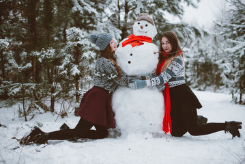 Close up fashion portrait of two sisters hugs and having fun, make snowman in winter time forest, wearing sweaters and scarfs,best friends couple outdoors, snowy weather