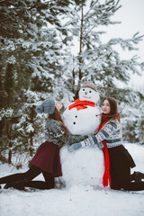 Close up fashion portrait of two sisters hugs and having fun, make snowman in winter time forest,...