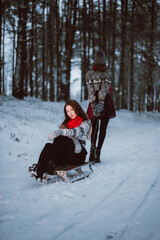 Close up fashion portrait of two sisters hugs and having fun, ride on sled in winter time forest, wearing sweaters and scarfs,best friends couple outdoors, snowy weather