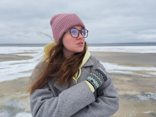 Portrait of beautiful young woman in hat, glasses and gloves  at winter cold day. Frozen girl freezing outdoors, walking