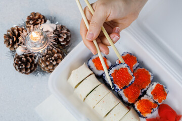 A set of Asian rolls in a plastic box at home in the kitchen in a festive style. The girl is holding food with chopsticks. Christmas concept. Decorations for the New Year