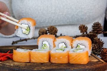 Japanese Philadelphia rolls on a wooden cutting board on a table in the kitchen. The girl is holding food with chopsticks. Christmas decoration. New Year concept