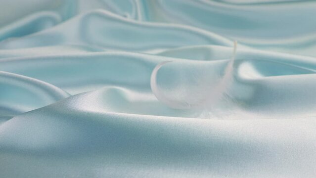 White swan feathers fall on pale blue pastel silk. Slow motion.