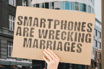 The phrase " Smartphones are wrecking marriages " is on a banner in men's hands with blurred background. Cyberspace. Media. Separation. Social. Upset. Mobile. Phone. Home. Bed. Conflict