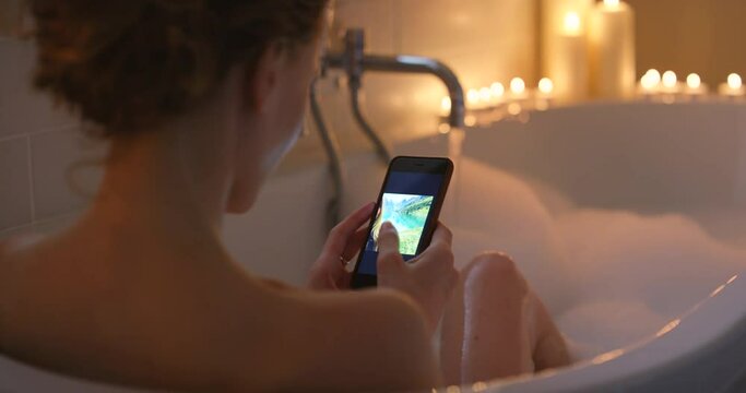 Woman, bathtub and using phone in bathroom for social media, photography to scroll and mobile app connection. Back view of girl enjoy bubble bath with smartphone, technology and browsing pictures