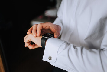 A man, a businessman in a white shirt holds a smartwatch, a wristwatch on his hand, preparing in the morning. Close-up photography, business.