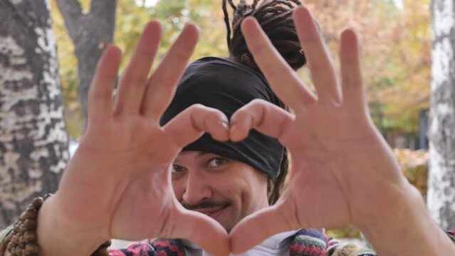 A young handsome man with dreadlocks and a beautiful smile made a heart sign with his hands looking through a heart from his palms smiling cute and cheerful. Positive guy with a beard
