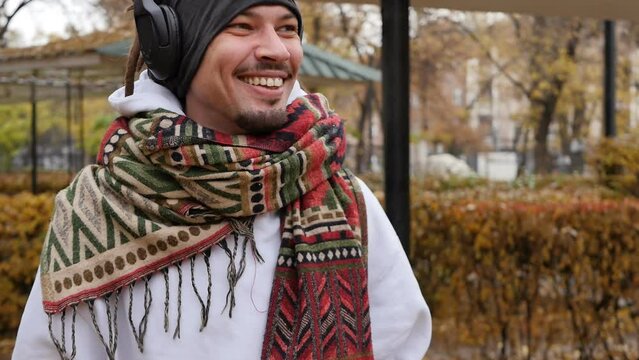 A handsome young mixed race guy with dreadlocks and a stylish beard wears a scarf around his neck and listens to his favorite music using headphones moving his head to the beats of the melody