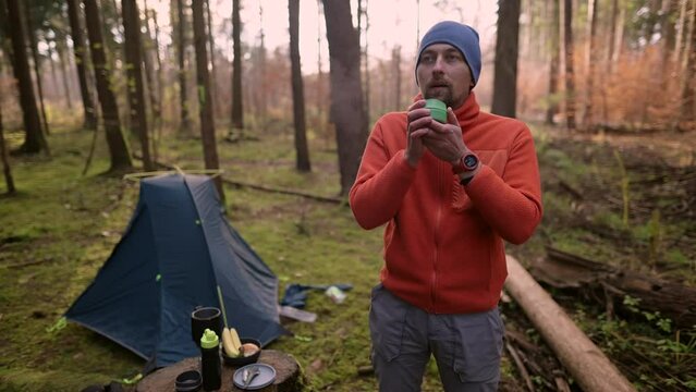 Man hiker camped in the woods and drinking tea from a teacup in front of the tent in the fall. Concept of traveling, hiking and trekking happy man having a cup in the campground autumn in the forest. 