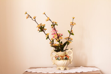 artificial flowers in a white ceramic vase on a knitted napkin on a wooden bedside table with a white background - close-up