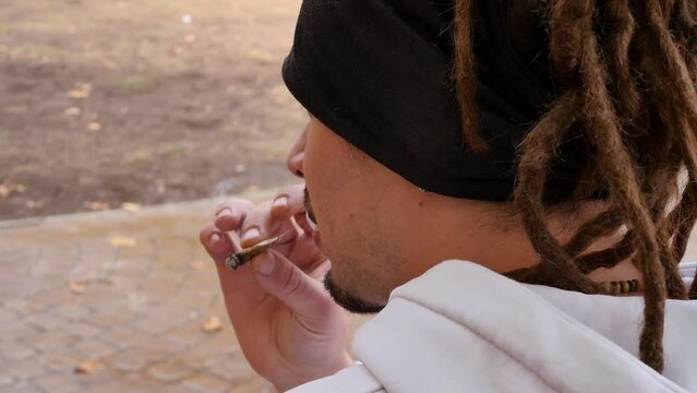 View from behind: a young guy with dreadlocks and a bandage on his hair in a white sweater smokes a cigarette with marijuana while sitting on a street bench. Legalized marijuana in some regions