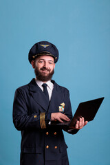 Smiling airliner pilot using laptop, typing message side view. Middle aged aviator in professional flight uniform holding portable computer in airport, browsing internet, online connection