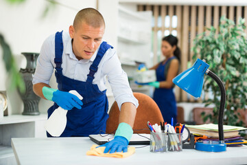 Portrait of professional worker of office cleaning service wiping furniture with rag and detergent ..