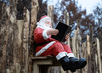 Bright toy Santa Claus sits on a wooden fence with a book in his hands