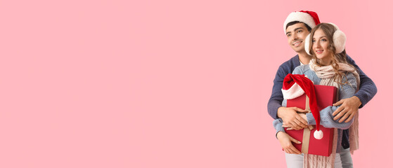 Young couple in love holding Christmas gift on pink background with space for text