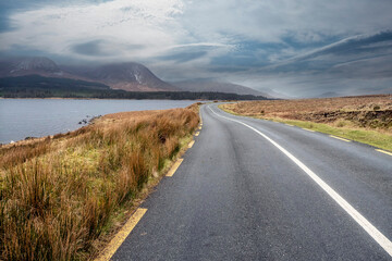 Beautiful scenery and small road by a lake in Connemara, Ireland. Mountains and cloudy sky in the background. Travel and transportation concept.
