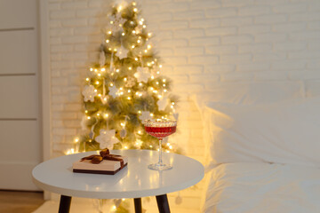 A glass of wine and a gift lie on the table against the background of a Christmas tree