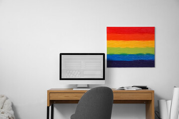 Modern workplace with computer and painting of LGBT flag hanging on light wall