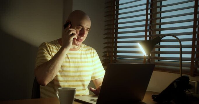 Man professiona works late at night, talking on the phone, solving remote work issues in a chat while working on a computer in the office. Smiling man is actively talking while sitting at a laptop.