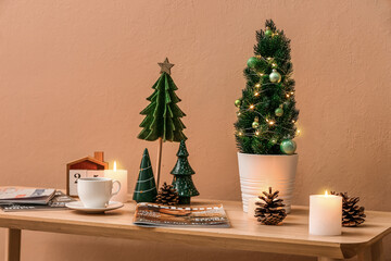 Small Christmas trees with candles, cup of coffee and magazines on table near beige wall