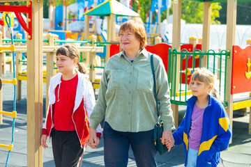 Mature smiling grandmother walks with her happy grandchildren on playground in summer, Russia