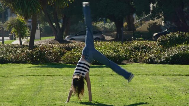 Young, fit woman turning a summersault along a green grassy meadow outdoors. Cheerful, athletic female making gymnastics movement in the park. High quality 4k footage