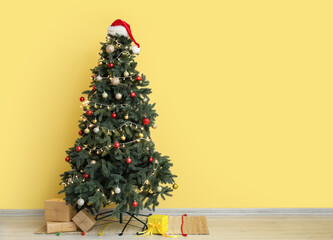 Glowing Christmas tree with Santa hat and presents near yellow wall