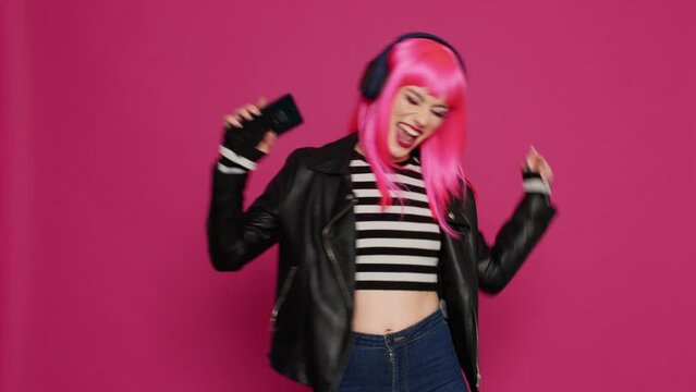 Happy beautiful girl with pink wig texting messages, listening to music on headset. Cool stylish woman having fun with mp3 songs, using social media online app on smartphone in studio.