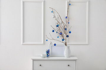 Vase with tree branches, Christmas balls and cup of coffee on drawers near light wall