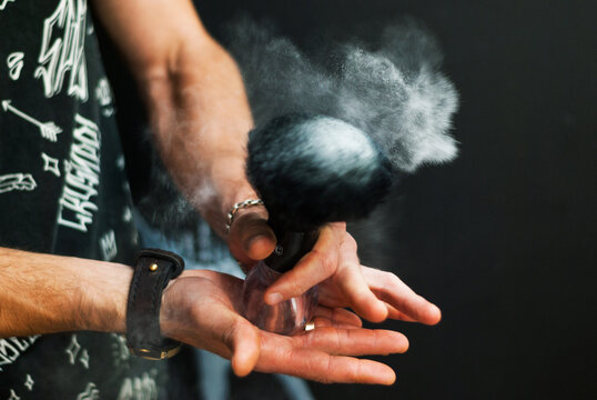 Men's hands hold a shaving brush from which talc scatters. Male stylist with shaving tools on a black background. Barber at work. Hair and beard care. Barbershop. © Sofiia