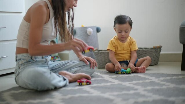 Little cute latin toddler playing with his car toys with his mom having fun sitting on the floor on a grey carpet wearing yellow T-shirt