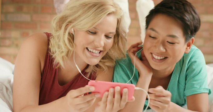 Couple, lgbtq and phone for streaming video online, comic fun or watching movie with earphones together in home. Lesbian women, happiness and smartphone for quality time or relax laughing on bed