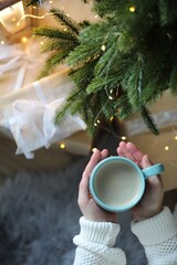 Woman holding cup of cocoa near Christmas tree indoors, top view