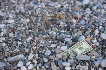 Hundred dollars half covered with round rocks lie on beach close up. Dollar bills partially buried...