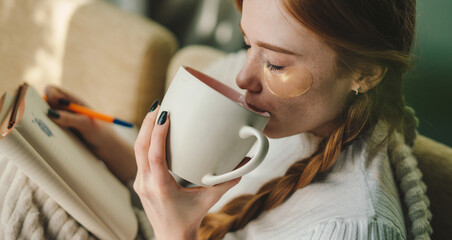 Pleasant young caucasian woman sitting on the sofa and drinking a mug of coffee wearing under-eye patches while writing future business plans in a diary