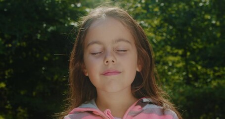 Portrait of a little girl child standing in the forest in the park. A preschool girl stands with her eyes closed and after a short time opens her eyes and looks at the camera.