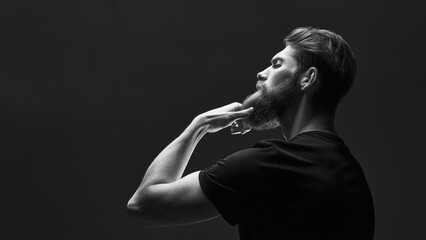 Black and white portrait of attractive pensive young man looks into the distance stroking his beard. Side view portrait of thinking stylish young man touch his beard.