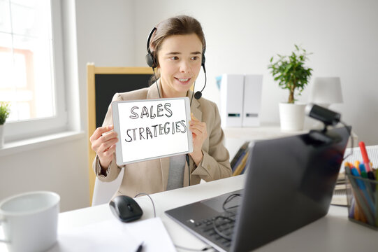 Young Woman Is Teaching English For Business With Laptop Computer, Camcorder And Headphone At Home. Teacher Is Having Video Conference Chat With Student. Person Hold Banner 