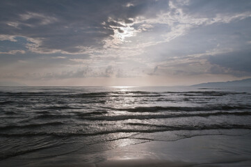 View from a beach looking toward the low sun partially hidden by clouds with waves breaking in foreground