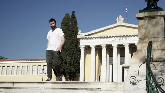 Bearded man walks in front of Athens Historic Zappeion sunny day in white shirt 