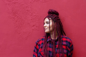 Portrait of young woman with red dreadlocks wearing a red checkered shirt standing by magenta wall...