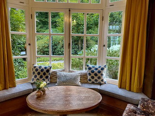 Wooden table near bay window seat with cushions in cozy room. Interior design - obrazy, fototapety, plakaty
