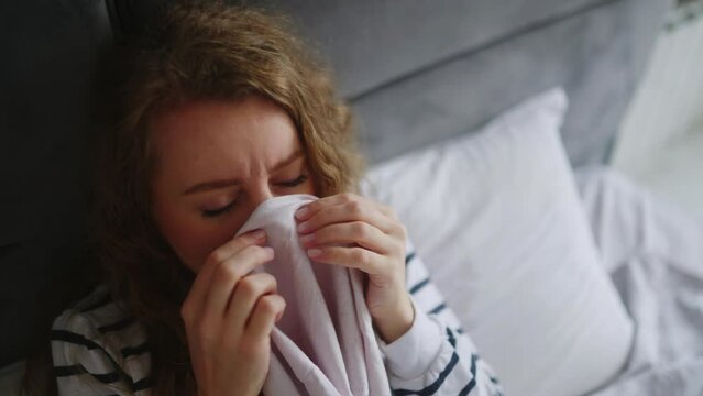 Woman sick with flu coughing and blowing her nose on her bed at home having sore throat, fever and headache. Young caucasian female suffering from coronavirus Covid19 while isolating touches forehead