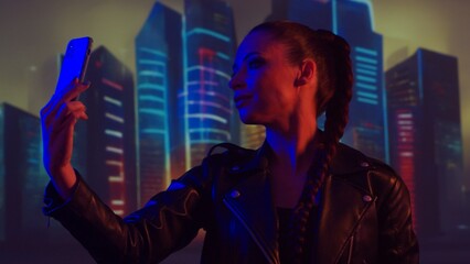Fototapeta premium Portrait of cyberpunk girl with a smartphone. Beautiful young woman on the background of city scyscrapers. Futuristic concept.