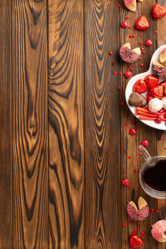Valentine's Day concept. Top view vertical photo of dish with confectionery cookies heart shaped candies and glass cup of drinking on wooden table background with empty space