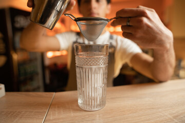 bartender holds in his hands sieve and steel shaker cup over glass with ice cubes.